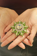 Hand holding a decorative brooch with green gemstones and gold accents.
