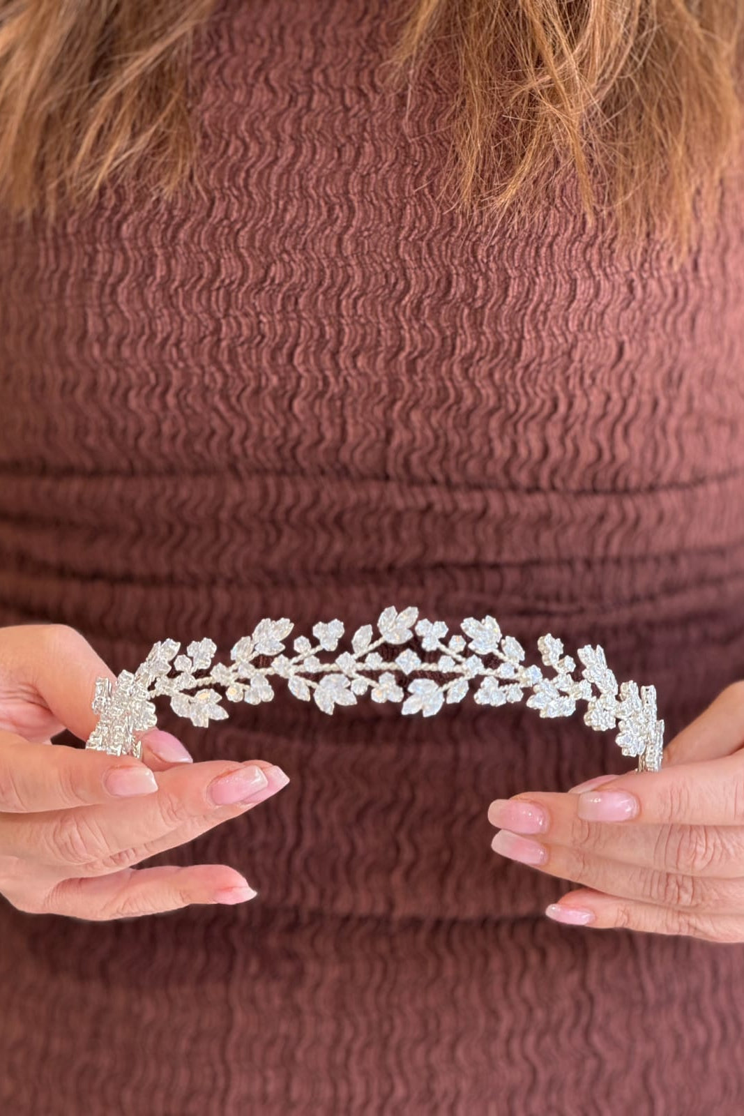 Close-up of a model holding a delicate, low-profile silver bridal headpiece featuring symmetrical branches of shimmering marquise crystal leaves meeting gracefully in the center.