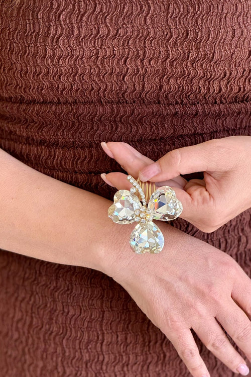 Close-up of a model holding a handcrafted gold-plated hair comb featuring a three-leaf clover design made of large, brilliant clear faceted crystals with a paved center.