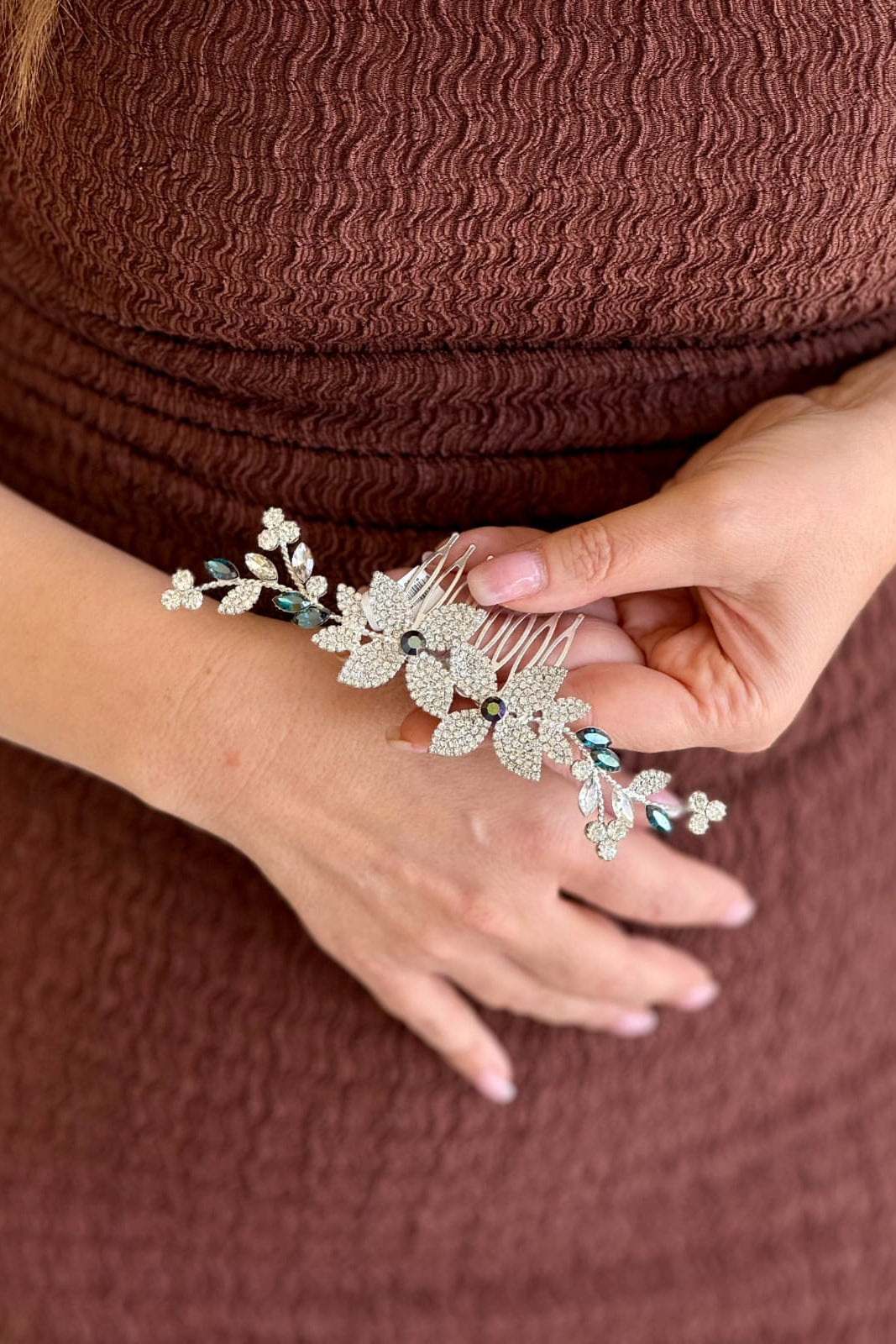 Close-up of a model holding a handcrafted silver-plated hair comb featuring an asymmetrical floral vine design set with clear pavé crystals and deep teal accents.